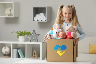 Humanitarian aid for Ukrainian refugees. Cute little girl holding donation box with toys indoors