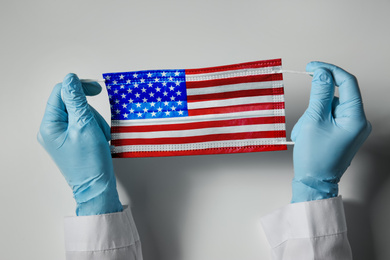 Doctor holding medical mask with USA flag pattern on light grey background, closeup. Dangerous virus