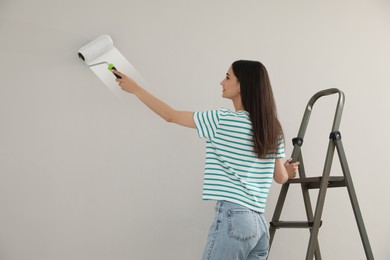 Photo of Young woman painting wall with roller on ladder indoors