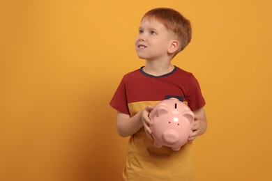 Photo of Cute little boy with ceramic piggy bank on orange background, space for text