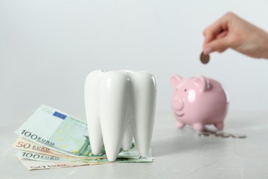 Woman putting coin into piggy bank at table, focus on ceramic model of tooth and euro banknotes. Expensive treatment