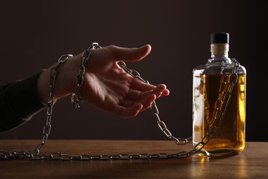 Alcohol addiction. Man chained with bottle of whiskey at wooden table, closeup