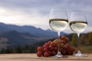 Photo of Glasses of tasty wine and grapes on wooden table against mountain landscape. Space for text
