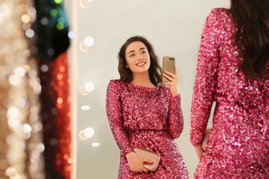 Photo of Beautiful young woman trying on stylish pink sequin dress and taking selfie near mirror in boutique. Party outfit