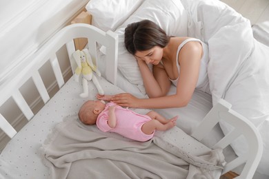 Young mother resting near crib with sleeping newborn baby at home, above view