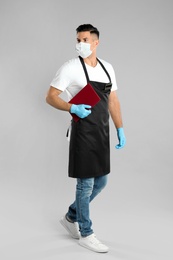 Waiter in medical face mask with menu on light grey background