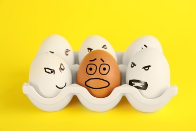 Photo of Brown egg with scared face among aggressively depicted white ones in holder on yellow background. Stop racism