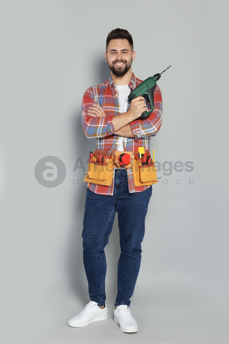 Young worker with power drill and tool belt on grey background