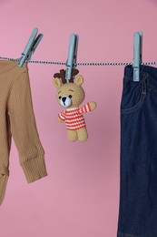 Different baby clothes and bear toy drying on laundry line against pink background
