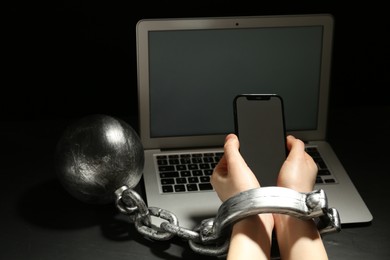 Woman shackled with ball and chain holding smartphone near laptop on dark background, closeup. Internet addiction