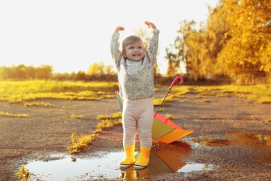 Photo of Little girl wearing rubber boots standing in puddle outdoors, space for text. Autumn walk