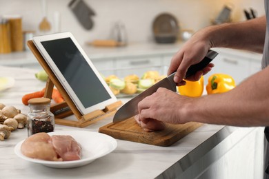 Man cutting chicken fillet while watching online cooking course via tablet in kitchen, closeup