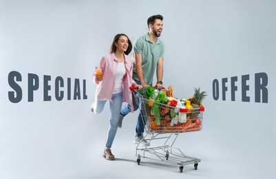 Special offer. Young couple with shopping cart full of groceries on grey background