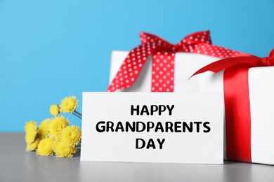 Card with phrase Happy Grandparents Day, gift boxes and beautiful flowers on grey table