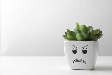 Photo of Beautiful potted houseplant with angry face on white table against light background, space for text. Emotional management