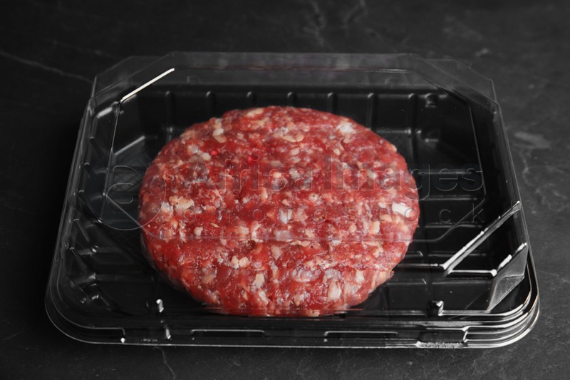 Plastic container with raw meat cutlet for burger on black table