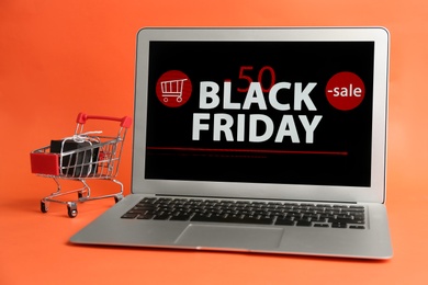 Laptop with Black Friday announcement, small shopping cart and gift on orange background