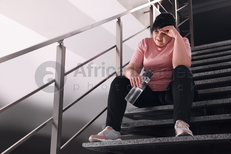 Photo of Tired overweight mature woman with bottle of water sitting on stairs indoors. Space for text