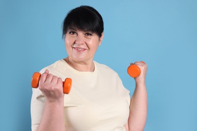 Happy overweight mature woman doing exercise with dumbbells on light blue background