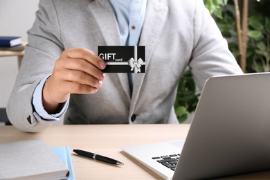 Man with gift card and laptop at table indoors, closeup