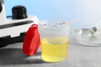 Container with urine sample for analysis and test strips on grey table in laboratory, closeup