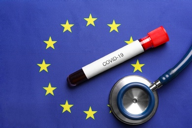 Stethoscope and test tube with blood sample on European Union flag background, flat lay. Coronavirus outbreak