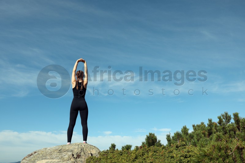 Beautiful young woman stretching on rock in mountains, back view