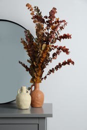 Vases, dried eucalyptus branches and stylish round mirror on grey table near white wall indoors. Interior design