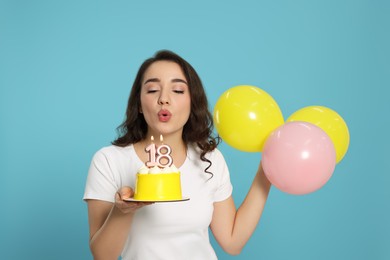 Photo of Coming of age party - 18th birthday. Woman blowing number shaped candles on cake and holding balloons against light blue background
