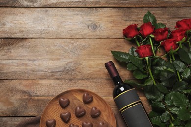Photo of Bottle of red wine, beautiful roses and heart shaped chocolate candies on wooden table, flat lay. Space for text