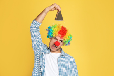 Funny man with clown nose, party hat and rainbow wig on yellow background. April fool's day