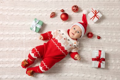 Cute little baby in Christmas outfit surrounded by festive items on white knitted plaid, top view. Winter holiday