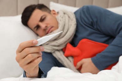 Photo of Ill man with hot water bottle checking his temperature at home