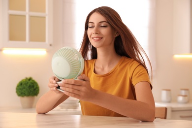 Woman enjoying air flow from portable fan at table in kitchen. Summer heat