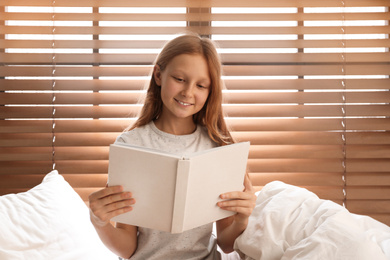Cute preteen girl reading book on bed near window