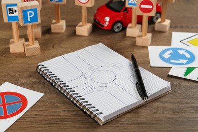 Many different road signs, notebook with sketch of roundabout and toy car on wooden table. Driving school