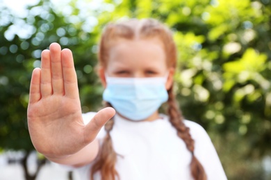 Little girl in protective mask showing stop gesture outdoors, focus on hand. Prevent spreading of coronavirus