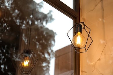 Pendant lamp with glowing light bulb near window indoors. Space for text