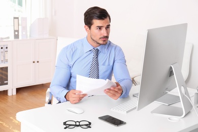 Handsome young man working with computer and papers at table in office