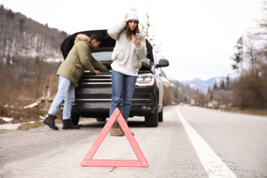 Photo of Man and woman near broken car outdoors, focus on emergency stop sign. Winter day