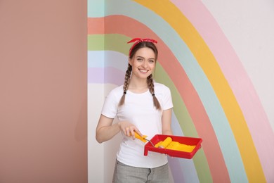 Young woman holding tray and roller near wall with painted rainbow indoors