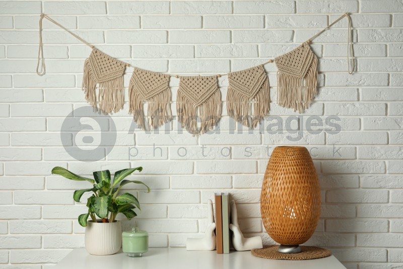Potted houseplant, decor elements and books on white table near brick wall with stylish macrame