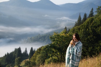Young woman with cup of hot drink and plaid enjoying beautiful mountain landscape. Space for text