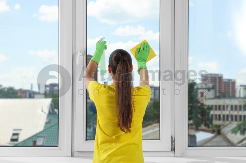 Photo of Woman cleaning window glass with sponge cloth and spray indoors, back view