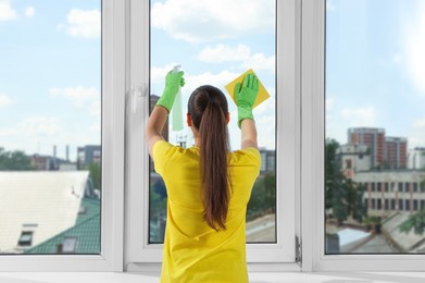 Woman cleaning window glass with sponge cloth and spray indoors, back view