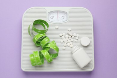 Scales with weight loss pills and measuring tape on violet background, top view
