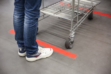 Person with shopping cart standing behind taped floor marking in store for social distance, closeup. Preventive measure during coronavirus pandemic