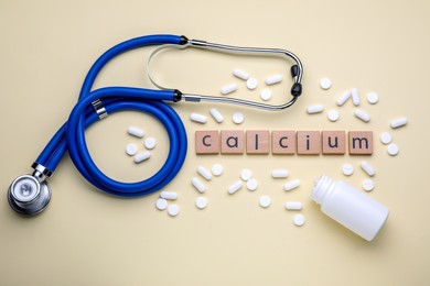 Photo of Word Calcium made of wooden cubes with letters, stethoscope and pills on beige background, flat lay