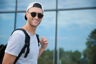Handsome young man with stylish sunglasses and backpack near reflection surface outdoors, space for text