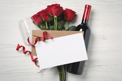 Flat lay composition with blank greeting card, wine and roses on white wooden table. Valentine's day celebration